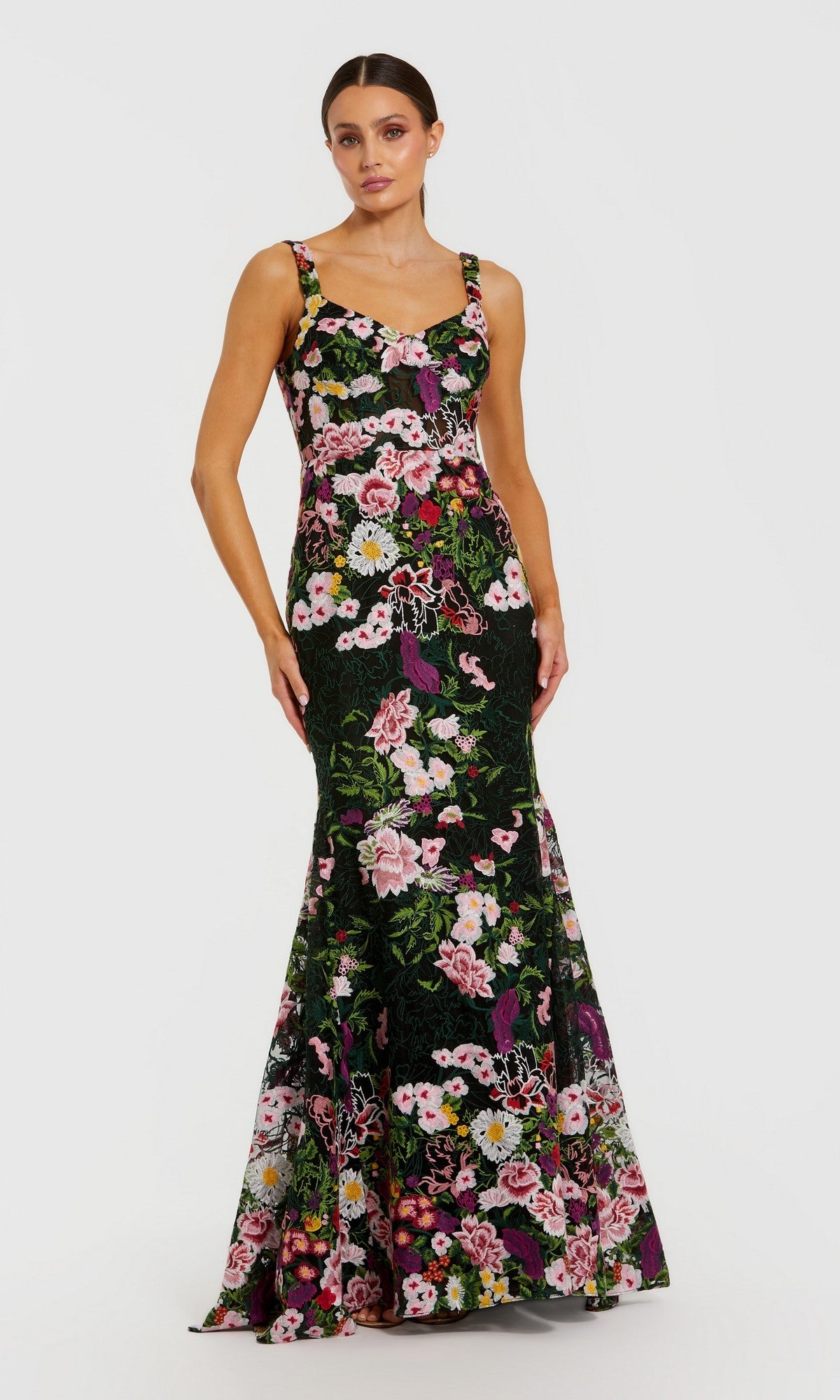 A woman stands looking forward with her arms relaxed by her sides, wearing the Mac Duggal 69004 long formal dress in Black Multi—a fitted, floor-length mermaid silhouette adorned with colorful floral embroidery.